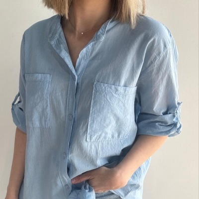 Blusa azul clara de manga comprida com bolsos frontais e botões, usada com calças de ganga claras.