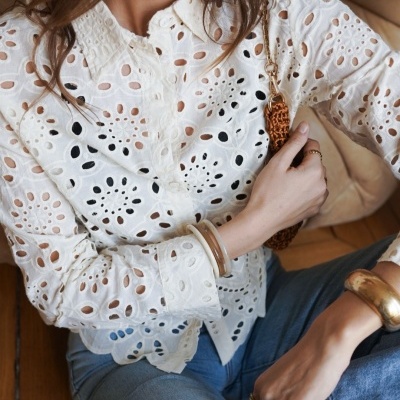 Blusa branca feminina bordada e recortada, jeans azul claro e acessórios dourados e castanhos.