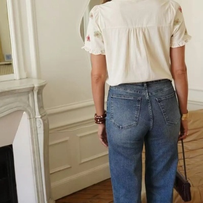 Pessoa de costas com blusa branca bordada, calças de ganga azuis e lenço vermelho em ambiente interior.