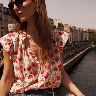 Mulher com blusa floral colorida, óculos de sol pretos, calças de ganga azuis, joias douradas, junto a um rio numa cidade.