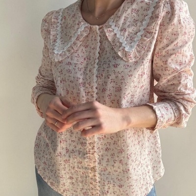 Camisa feminina bege com flores cor-de-rosa e gola com folhos e fita branca ondulada.