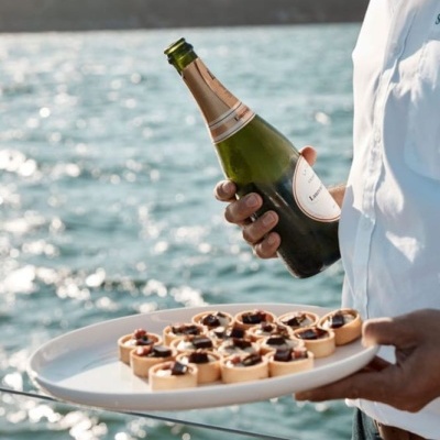 Pessoa com camisa branca segura garrafa de champanhe e prato com mini tartes junto ao mar