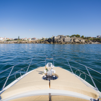 Proa de barco com almofadões bege no mar em frente a costa rochosa com edifícios