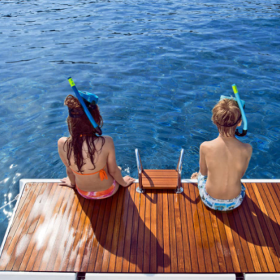 Duas crianças com máscaras de mergulho sentadas em plataforma de madeira de barco no mar azul