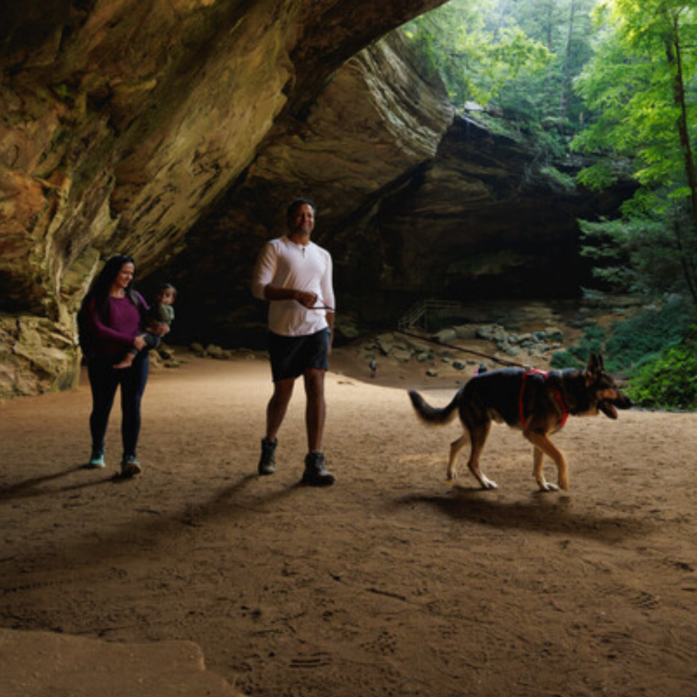 Front Range® Dog Harness - Red Canyon - Ruffwear Cão Pastor Alemão com peitoral vermelho e família a caminhar em caverna com chão de terra