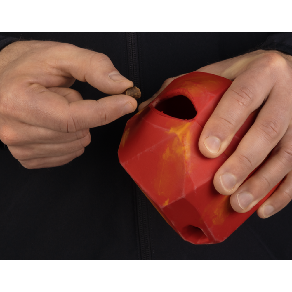 Gnawt-a-Rock™ - Glacial Blue Brinquedo para cães vermelho em borracha com orifício para petisco, mãos segurando o brinquedo e um petisco pequeno