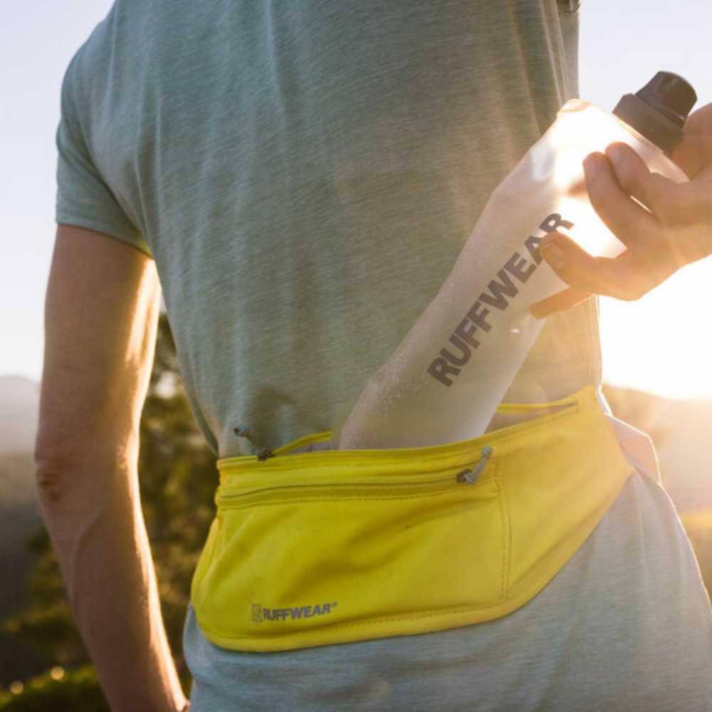 Trail Runner™ Running Belt - Lichen Green Pessoa com camiseta cinza e pochete amarela colocando garrafa RUFFWEAR ao ar livre
