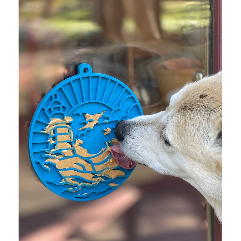 Tapete de lamber Lighthouse eMat! LickMat Decoração azul com relevo dourado de navio e gaivotas na superfície de vidro com cão branco a lamber.