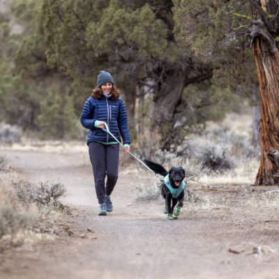 Mulher com casaco azul caminha com cão preto com colete e botas verdes numa trilha