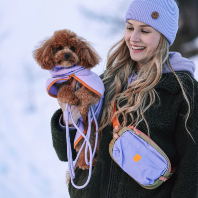Mulher segura cão de casaco lilás, usando gorro lilás e bolsa lilás no peito, ambiente com neve.