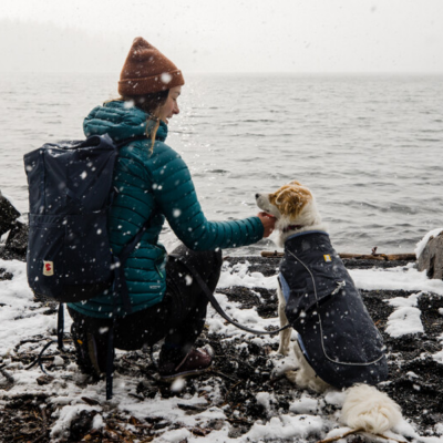 Mulher de casaco azul acolchoado e gorro castanho com mochila azul e cão com casaco preto na neve junto ao mar