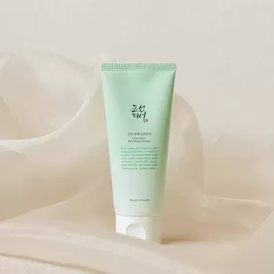 Tubo verde claro de Green Plum Refreshing Cleanser com tampa branca sobre tecido creme