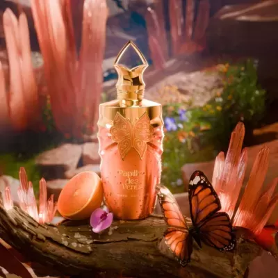 Frasco de perfume cor de pêssego e dourado com borboleta decorativa, sobre tronco com borboletas e cristais cor de rosa