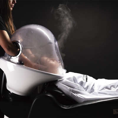 Aparelho facial com cúpula transparente e vapor, pessoa deitada com roupa branca.