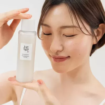 Frasco translúcido de cosmético Glow Replenishing Rice Milk segurado por mulher com cabelo castanho