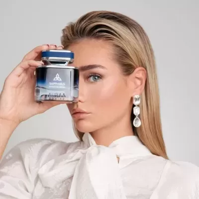 Frasco de perfume azul e transparente segurado por mulher loira de blusa branca