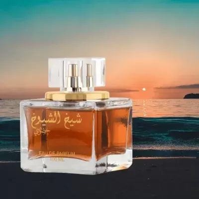 Frasco de perfume quadrado com líquido âmbar, detalhes dourados e texto em árabe, sobre praia ao pôr do sol