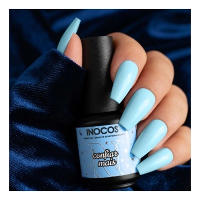 Unhas compridas pintadas de azul claro segurando frasco de verniz INOCOS