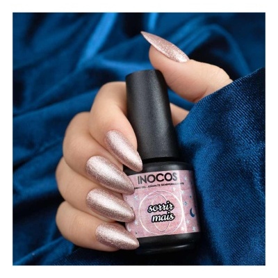 Verniz para unhas INOCOS com encaixe preto e etiqueta rosa, unhas pintadas rosa brilhante seguras por mãos sobre tecido azul