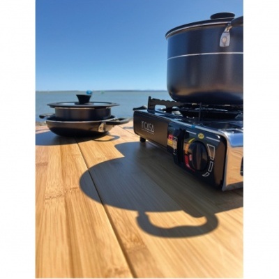 Fogão portátil com panela e frigideira pretos sobre mesa de madeira com mar ao fundo