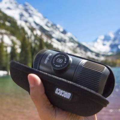 Monocular preto em capa preta, mão a segurar, fundo natural com lago e montanhas nevadas
