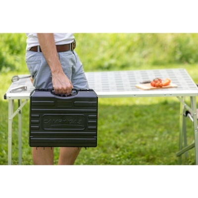 Mala preta rígida com o texto CAMPINGAZ, segurada por pessoa junto a mesa dobrável exterior