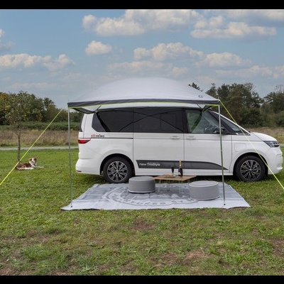 Caravana branca com toldo estendido, tapete cinza, dois pufes e mesa pequena num campo verde sob céu azul com nuvens