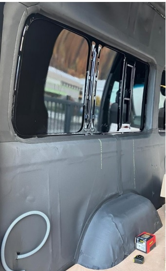 Interior de carrinha com paredes cinzentas, janela escurecida, tubo branco, caixa vermelha e preta e fita métrica no chão