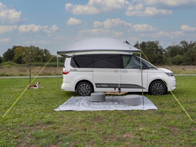 Caravana branca com toldo estendido, tapete cinza, dois pufes e mesa pequena num campo verde sob céu azul com nuvens