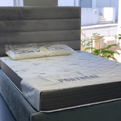 Cama de casal com colchão branco, estrutura cinza e duas almofadas embaladas