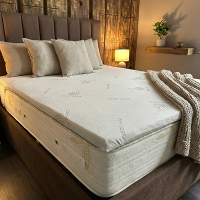 Cama estofada castanha com colchão branco estampado aloé vera, cinco almofadas bege, cobertor grosso bege, abajur e planta em decoração de quarto rústico