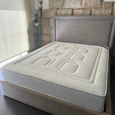 Colchão branco geométrico sobre base de cama estofada bege e cabeceira cinza em ambiente com parede de pedra e candeeiro