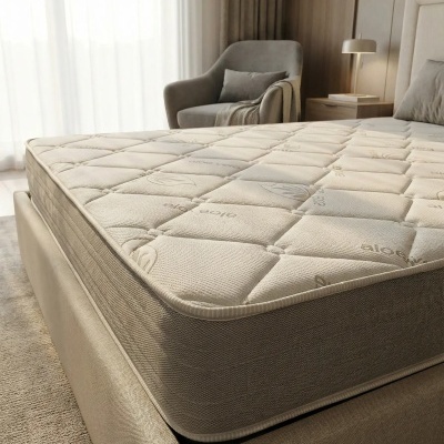 Colchão branco com padrão e texto aloe vera em cama estofada bege, cadeira cinza ao fundo