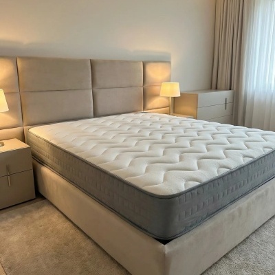 Cama estofada cinza claro com colchão branco, candeeiros e cortinas claras