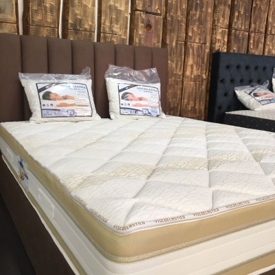 Cama estofada castanha com colchão branco e bege e almofadas embaladas em plástico com parede de madeira ao fundo.