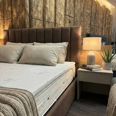 Cama estofada castanha com cabeceira acolchoada, colchão com padrão aloe vera, almofadas e manta bege, mesa de cabeceira com candeeiro e planta, parede de madeira rústica.