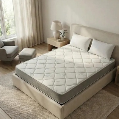 Cama de casal com colchão branco e almofadas brancas numa sala com mesa de cabeceira, candeeiro e cortinas translúcidas