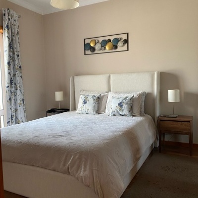 Quarto com cama dupla estofada branca, roupa de cama bege clara, almofadas florais, mesas de cabeceira em madeira com candeeiros, cortinas florais e quadro colorido na parede.