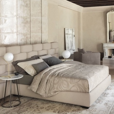 Quarto com cama de casal estofada bege, mesa de cabeceira com candeeiro branco, tapete clássico e lareira com espelho