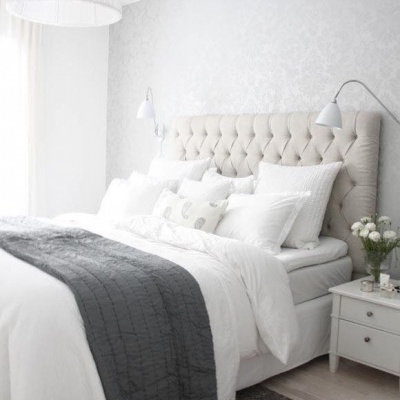 Quarto com cama estofada bege, almofadas brancas, manta cinza e mesa de cabeceira com flores