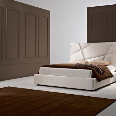 Quarto moderno com cama estofada branca e cabeceira geométrica, roupa de cama castanha, tapete castanho, mesa de cabeceira branca com candeeiro e armário castanho.