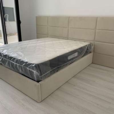 Cama de casal com cabeceira estofada em tecido bege e colchão embalado em plástico numa sala clara