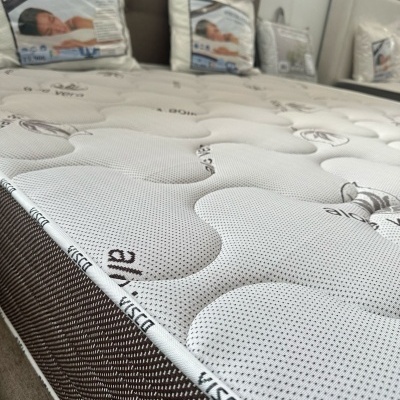 Colchão viscoelástico branco com padrão ondulado e faixa lateral castanha, com almofadas na embalagem ao fundo