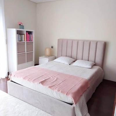 Quarto com cama de casal estofada cinza claro, cobertor rosa e estante branca com caixas e livros.