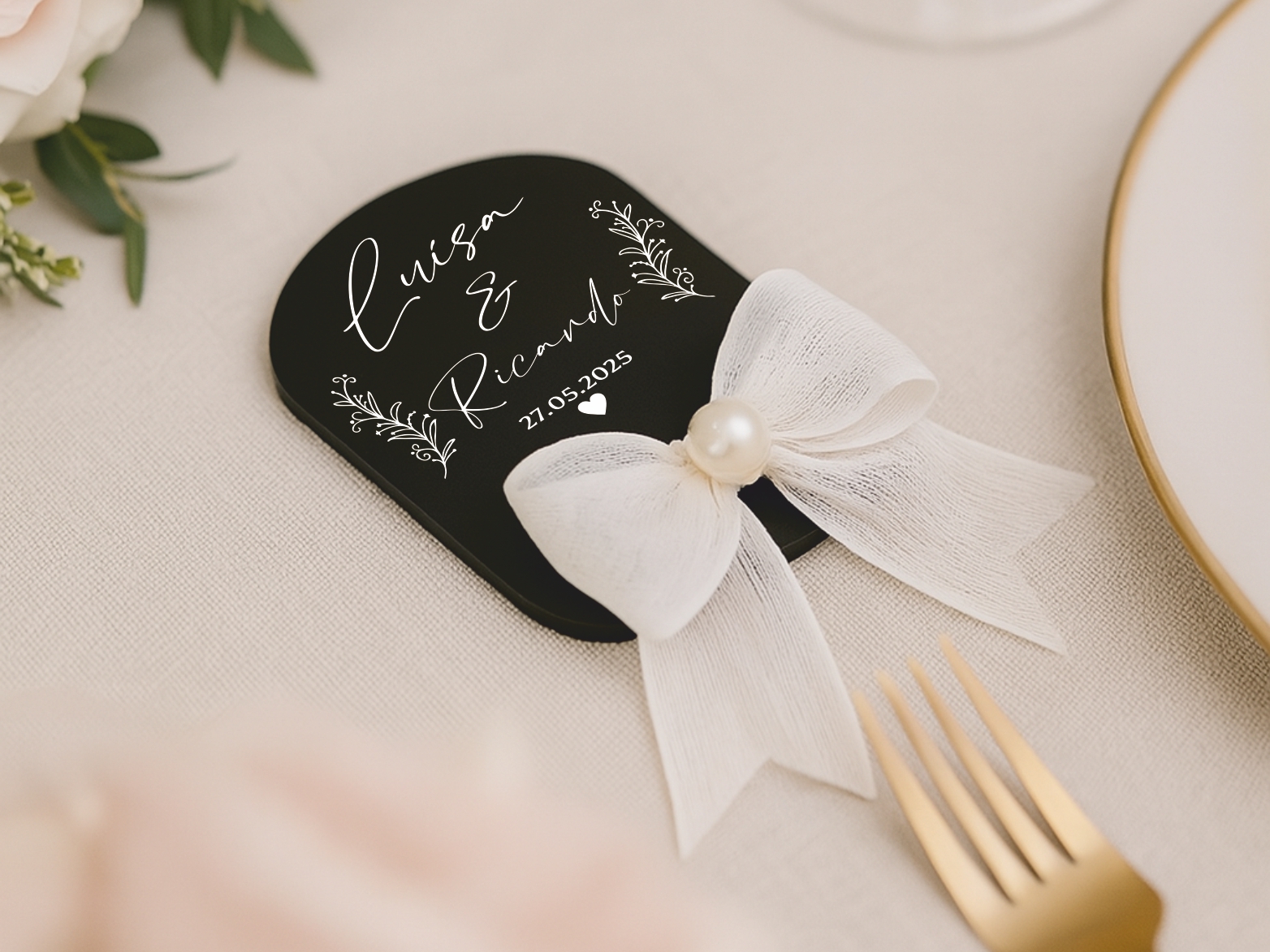 Lembrança de Casamento em Acrílico Preto com Laço – Modelo M2 Cartão decorativo preto com texto branco e laço branco com pérola, em cima de tecido bege, ao lado de talheres dourados.