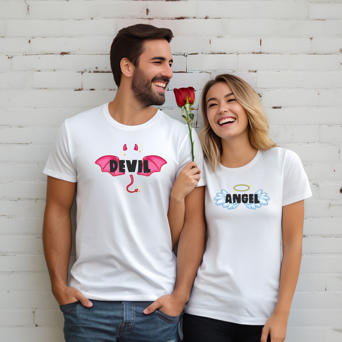 Casal com t-shirts brancas estampadas com DEVIL e ANGEL, segurando rosa vermelha em fundo de parede de tijolos brancos.