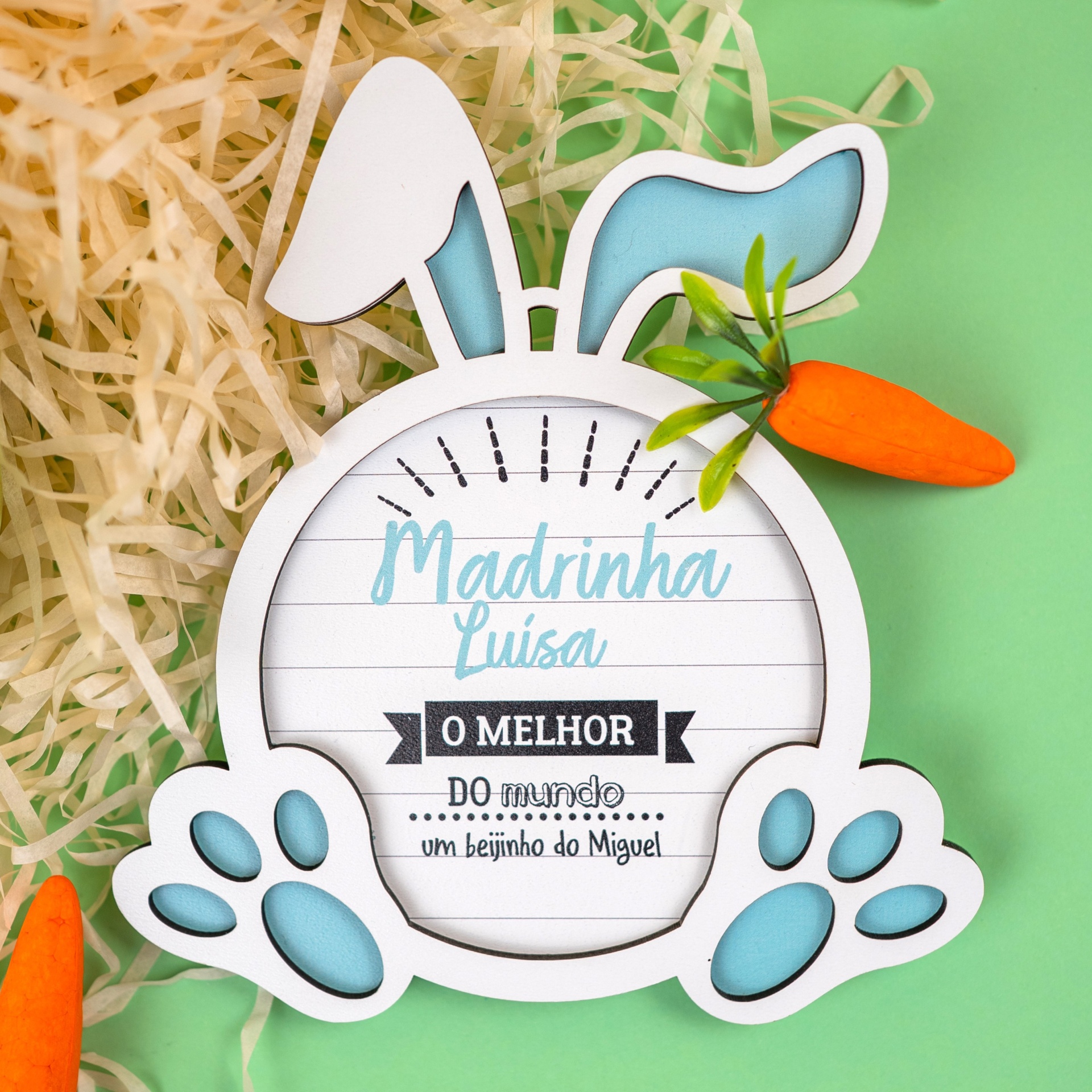Placa Coelhinho "Padrinho/Madrinha o melhor..." Placa decorativa de coelho branca com orelhas e patas azuis e texto para madrinha, cenoura de plástico e palha amarela, fundo verde.