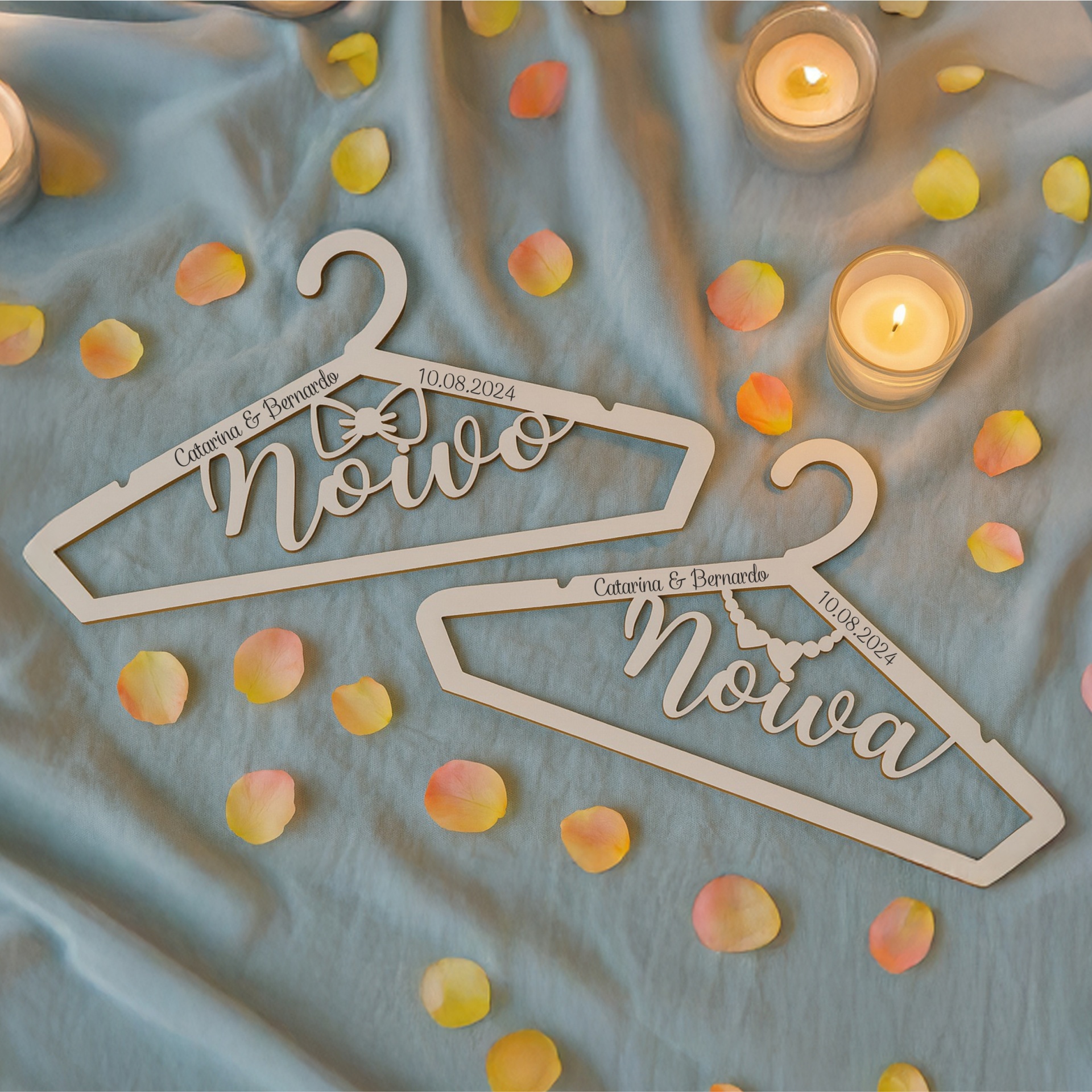 Conjunto de Cruzetas Personalizadas Noivo & Noiva – Modelo 1 Cabides de madeira branca personalizados com nomes e data sobre tecido azul com pétalas e velas