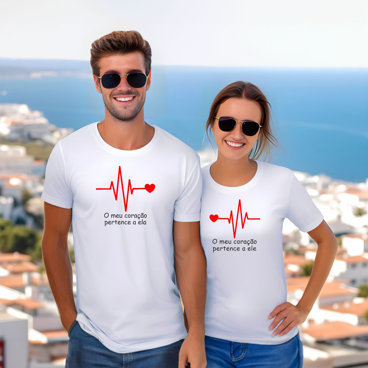 Casal com t-shirts brancas pintadas com linha de batimento cardíaco vermelha e texto romântico, usando óculos escuros, com vista para o mar e edifícios ao fundo.