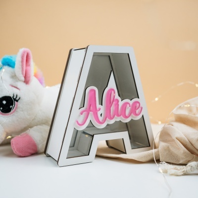 Letra A luminosa branca com texto Alice rosa junto de peluche de unicórnio sobre superfície branca